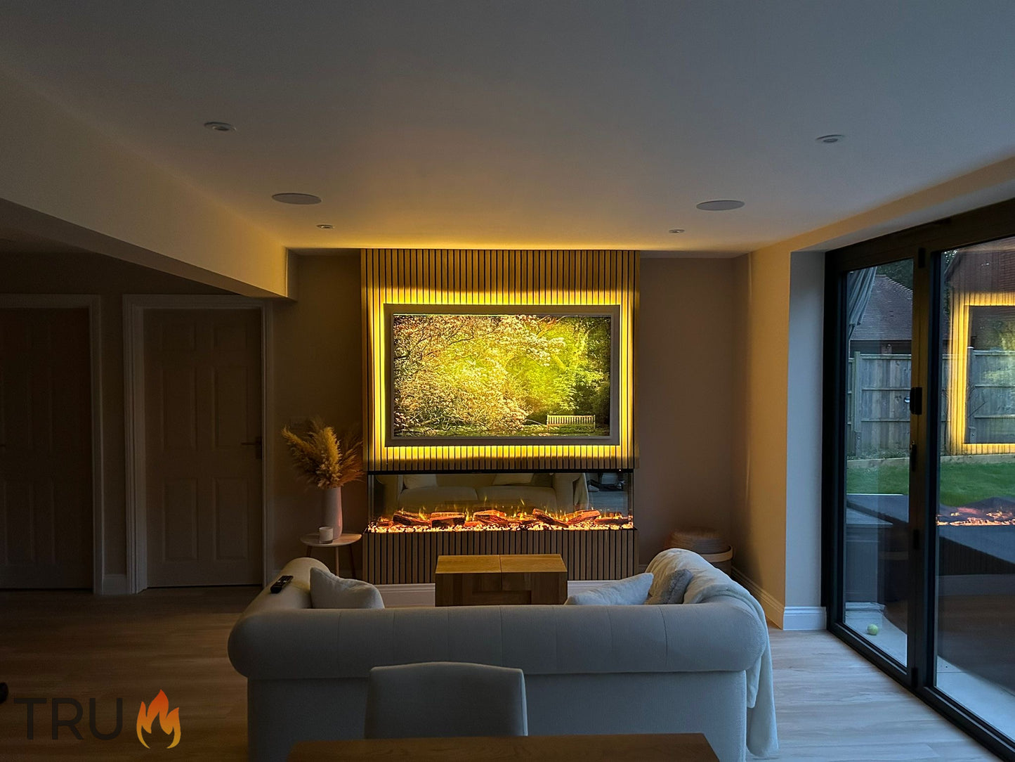 A modern 3-sided media wall electric fire with a glowing LED flame effect, mounted on a wall with a room with a white sofa in the foreground.